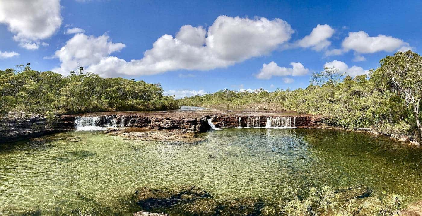 A wide, shallow waterfall pouring over a rock ledge into a clear pool surrounded by forest